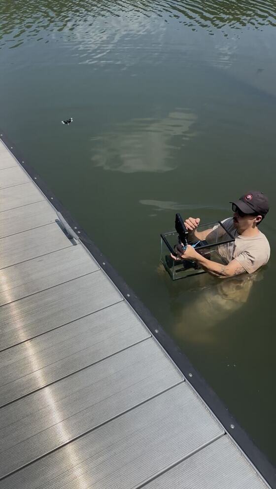 Jake waist-deep in a lake beside a dock, filming with a cinema camera rig